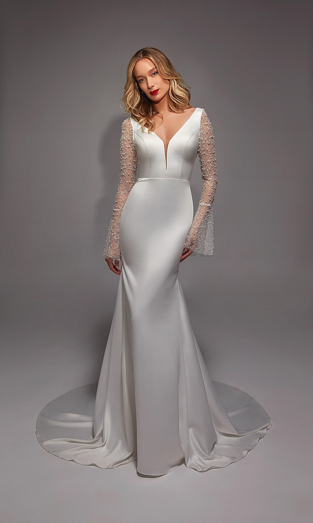 A woman wearing the Alyce 7137 long white dress, featuring satin fabric, illusion lace sleeves, a deep V-neckline, and a train, stands against a plain gray background.